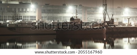 Cargo port terminal and river at night. Ship's, cranes, warehouse. Illuminated christmas tree, lights in the background. Concept dark urban scene. Symmetry reflections on the water