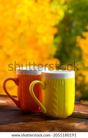 Yellow and orange ceramic cups with a knitted pattern on a background of yellow trees on a wooden table. autumn template and postcard. Warm and cozy autumn. Leaf fall
