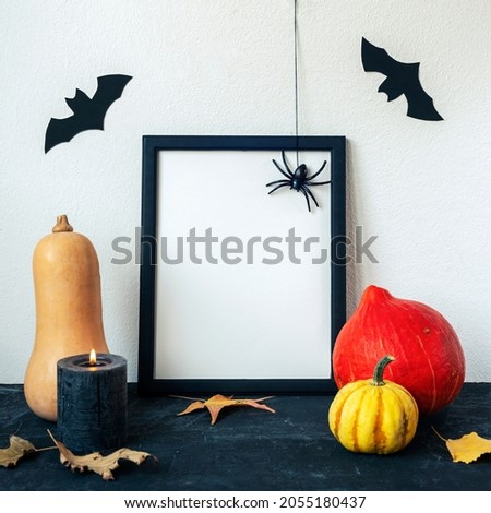 Halloween holiday concept with blank picture frame, pumpkins, candle, bats and spider on black table against white wall. Copy space, mock up.