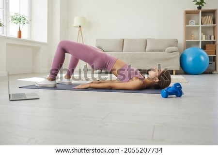 Fit woman doing workout at home. Young lady watching online video lesson on notebook PC, having virtual class with remote instructor, doing strengthening glute bridge gymnastic exercise on fitness mat Royalty-Free Stock Photo #2055207734