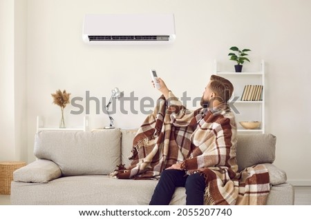 Sad angry man sitting on sofa trying to warm up under plaid while freezing in cold living room with modern air conditioner turned on. Young guy having trouble adjusting AC system temperature at home Royalty-Free Stock Photo #2055207740