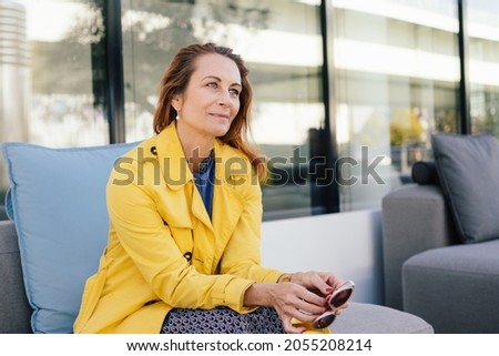 Elegant modern redhead woman in bright yellow jacket sitting waiting in a vestibule or waiting room holding her sunglasses looking ahead with a quiet smile and contemplative expression Royalty-Free Stock Photo #2055208214