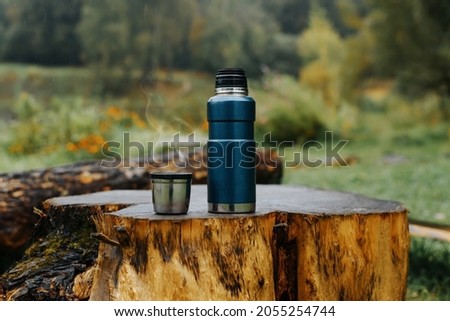 Thermos and aluminum hot drink mug with rising steam outdoors. Camping vacuum flask and iron cup standing on tree stump in rainy, cold weather. Hike, camping concept. Royalty-Free Stock Photo #2055254744