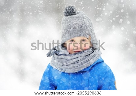 Portrait of funny little boy in blue winter clothes walks during a snowfall. Cute child wearing a warm clothing, hat, scarf is looking at you. Outdoors winter activities for family with kids.