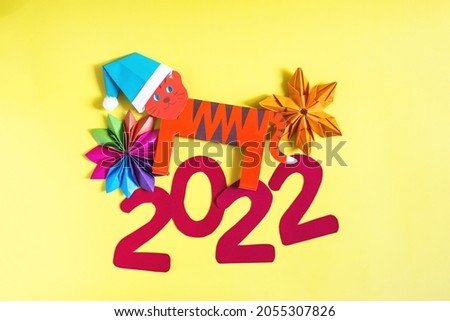 A colorful paper tiger in a Christmas hat and the numbers 2022 on a bright yellow background. The symbol of the new year is a paper tiger.