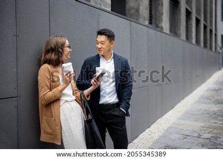 Asian businessman and caucasian businesswoman drinking coffee and talking in city. Concept of business cooperation. Idea of freelance and remote work. Woman with smartphone. Modern successful people