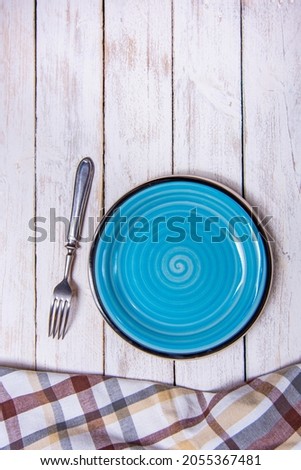 Top view background with empty blue ceramic plate, antique silver cutlery on vintage weathered white wooden boards. Copyspace. Brown checkered kitchen towel. Vertical photo.