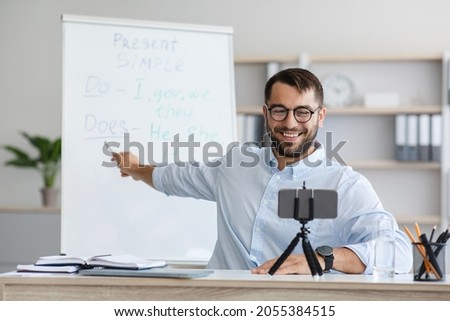Cheerful adult european male teacher with beard in glasses shows to blackboard and have video call with online class at home interior. Tutor, modern distance education and lecture due new normal Royalty-Free Stock Photo #2055384515
