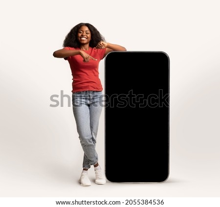 Cheerful Young African American Lady Leaning And Pointing At Big Smartphone With Blank Screen, Smiling Black Lady Showing Copy Space For Advertisement, Standing Over White Studio Background, Mockup Royalty-Free Stock Photo #2055384536