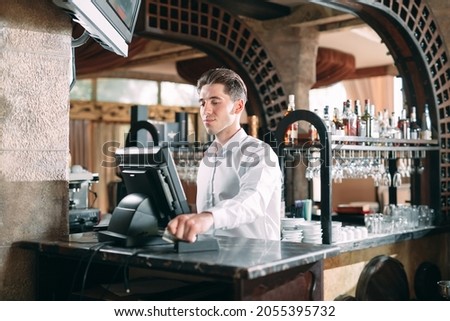 small business, people and service concept - happy man or waiter in apron at counter with cashbox working at bar or coffee shop