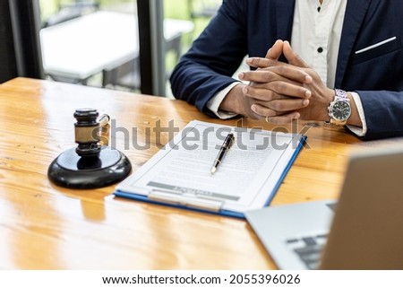 A male lawyer sits in his office, on a table with a small hammer to beat the judge's desk in court. and justice scales, lawyers are drafting a contract for the client to use with the defendant to sign Royalty-Free Stock Photo #2055396026