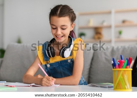 Lifestyle Concept. Smiling school girl with colorful dyed hair sitting at desk, writing essay in copybook. Pupil with headset studying and doing homework or taking notes in her diary, making checklist Royalty-Free Stock Photo #2055410462