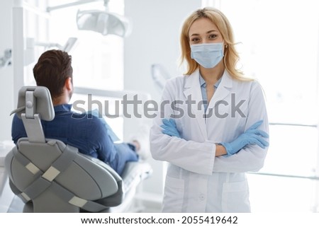 Female dentist blonde woman in protective face mask and medical gloves posing at modern dental clinic, unrecognizable man patient sitting in dental chair, dental care while coronavirus epidemic Royalty-Free Stock Photo #2055419642