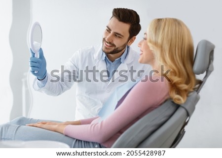 Friendly doctor dentist holding mirror for attractive young blonde woman patient sitting in dental chair, doctor and patient checking results of dental treatment, looking at mirror and smiling Royalty-Free Stock Photo #2055428987