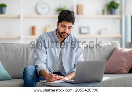 Arab male freelancer writing in notepad and using laptop, taking notes and using computer while sitting on sofa, noting information from internet, copy space