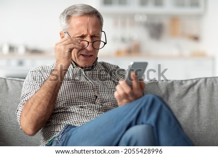 Poor Eyesight. Senior Man Squinting Eyes Reading Message On Phone Wearing Eyeglasses Having Problems With Vision Sitting On Couch At Home. Ophtalmic Issue, Bad Sight In Older Age Concept Royalty-Free Stock Photo #2055428996
