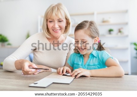 Loving smiling mature woman sitting at table with little girl engaged in game assemble jigsaw together, happy grandmother playing having fun with preschooler kid making picture connect puzzle pieces