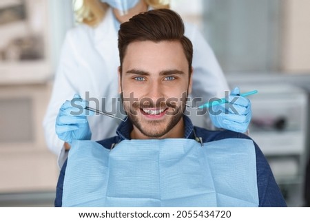 Smiling young bearded man sitting in dental chair, visiting modern dental clinic, having regular checkup, unrecognizable woman dentist holding dental tools next to male patient mouth Royalty-Free Stock Photo #2055434720