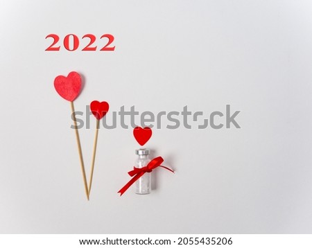 2022 and Vaccination card. Bottle of the coronavirus vaccine Covid-19 as a gift for Christmas, with a red bow and two hearts on wooden sticks on a white background with empty place for your text.