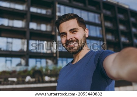 Close up young smiling sporty strong fit tourist sportsman man wear blue casual t-shirt warm up training do selfie shot on mobile cell phone outdoors near building in city centre in summer day morning