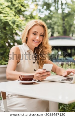 Vertical photo of successful mature businesswoman working remotely on distance on laptop, using smart phone in cafe. E-banking, e-commerce concept