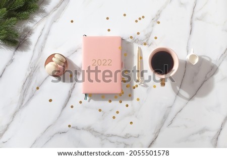 Flat lay composition with coral colored 2022 diary book for writing down New Year's plans, coffee, cookie and mini artificial Christmas trees onside with gold sparkles on white background. Holiday