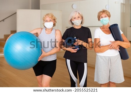 Three smiling aged women in sportswear and protective masks standing in fitness studio with sports equipment in hands, ready for training. Healthy lifestyle and pandemic precautions concept