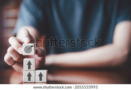 Businessman holding wooden cube with target board icon and arrow on wooden table. Goals and planning for success in marketing business, achieve the objective concept. Closeup and free copy space Royalty-Free Stock Photo #2055533993