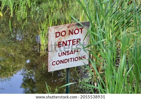A small, square, metal sign with red, bold, font that reads "Do not enter unsafe conditions". The sign is attached to a metal stake that is sitting in a body of water and hiding behind blades of grass