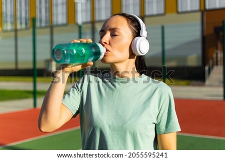Photo of a middle-aged woman in white headphones after workout drinking water from a plastic bottle.