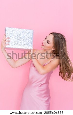 Image of beautiful young woman shaking box with gift wonder what inside wrapped box stands on pink background. Looking on the boxsur. Celebration concept. Happy birthday. New Year. 8 March 
