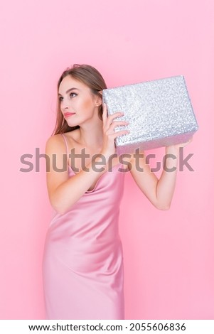Image of cute beautiful girl shaking box with gift wonder what inside wrapped box stands on pink background. Looking away. Celebration concept. Happy birthday. New Year. 8 March 