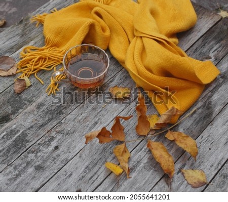          An orange scarf on a wooden background, a cup of tea and autumn leaves on a gray background.                     