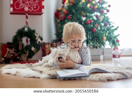 Beautiful toddler child, reading book with pet dog in front of  Christmas tree, decoration and presents around him