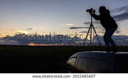 A girl photographs the sunset from a tripod, standing on the roof of a car in the field, copy space.