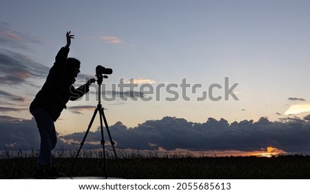 A girl photographs the sunset from a tripod, standing on the roof of a car in the field, copy space.