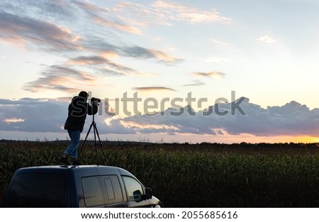 A girl photographs the sunset from a tripod, standing on the roof of a car in the field.