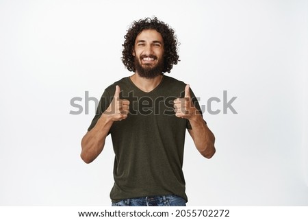 Enthusiastic handsome man smiling, showing thumbs up, approve and like, standing in casual clothes over white background