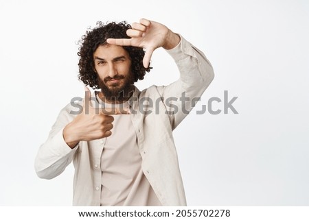 Handsome creative man looking through hand frames, searching perfect angle for photo shot, standing in casual clothes over white background
