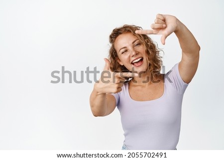 Happy blond girl looking through hand frames with joyful smile, picturing moment, taking picture gesture, standing over white background