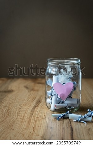 A mason jar with wish lists and a pink heart on a brown wooden table. Black background, close-up. 