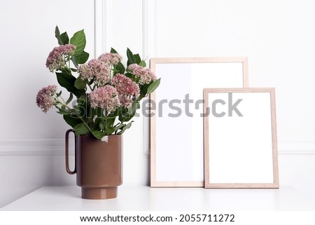 Stylish ceramic vase with beautiful flowers and blank frames on table near white wall