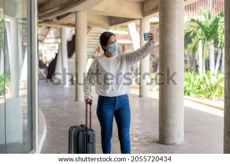 Travel. A woman in a white long-sleeved shirt and jeans, wearing a mask. Using a mobile phone for taking a selfie with suitcase on the right hand.