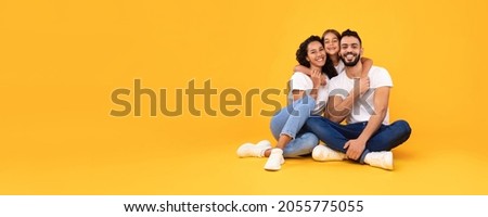 Arab Family Of Three Posing Hugging And Smiling To Camera Sitting On Yellow Studio Background. Panoramic Shot Of Young Arabic Parents Embracing Little Daughter. Empty Space For Text