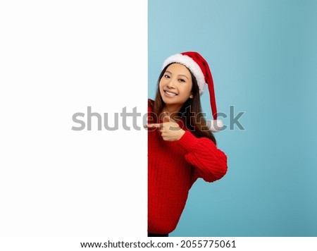 Christmas promo. Asian lady in Santa hat pointing at white empty banner with space for your ad over blue background, mockup for your design