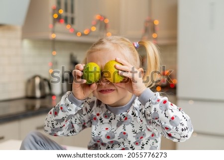 cute, joyful little blonde girl in Christmas print clothes is sitting in beautiful kitchen with two tangerines in her hands. lifestyle. new Year's breakfast. new year's winter concept. space for text