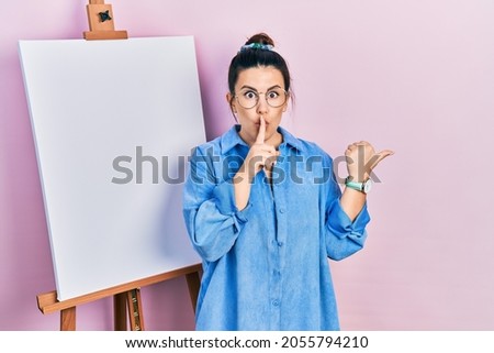 Young hispanic woman standing by painter easel stand asking to be quiet with finger on lips pointing with hand to the side. silence and secret concept. 