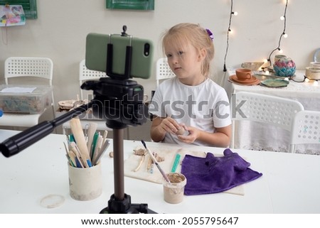 Girl vlogger recording video with modelling clay and speaking for smartphone camera. Kid blogger in a ceramic studio creating interesting content for children. 