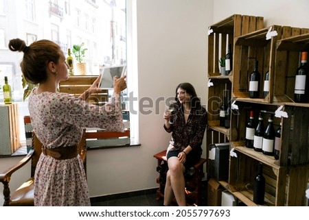 two female wine lovers take picture selfie with a tablet in a wine shop - bloggers vloggers posting on social networks during wine tasting - selective focus on background