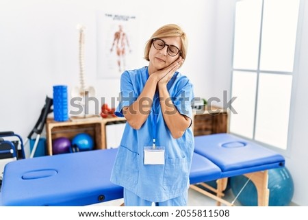 Middle age blonde woman working at pain recovery clinic sleeping tired dreaming and posing with hands together while smiling with closed eyes.  Royalty-Free Stock Photo #2055811508